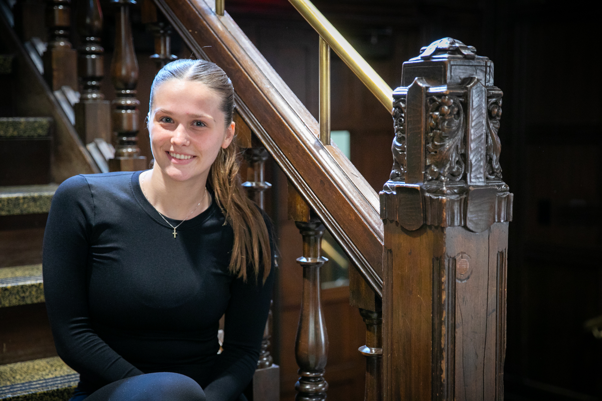 Lucy Rupertus sits on stairs next to an ornate wooden carved banister inside the ARCH building on Penn's campus.