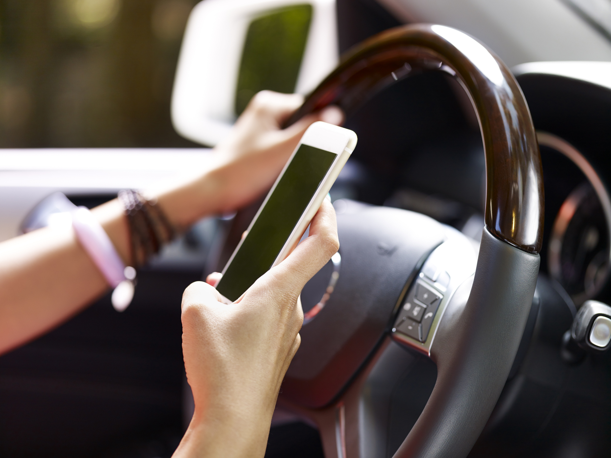 A teenagers hands holding a steering wheel in one hand and a smartphone in the other.