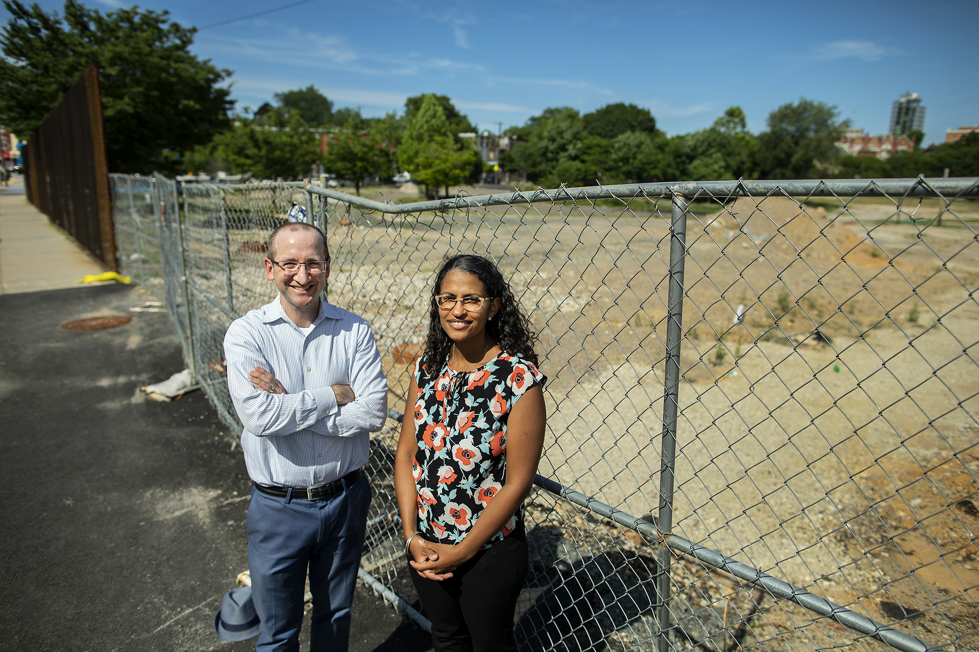 Two people standing in front of a fenced-in vacant lot, one leaning against the fence, the other standing with arms crossed.