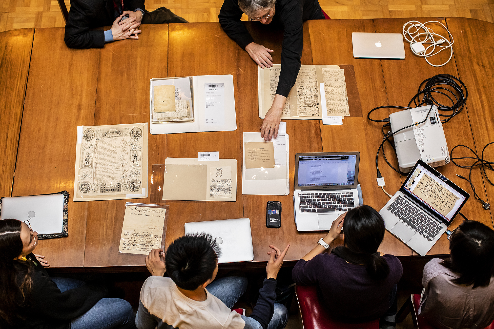 Overhead shot of rare documents spread out on wood table with students and professors and laptops surrounding them. 