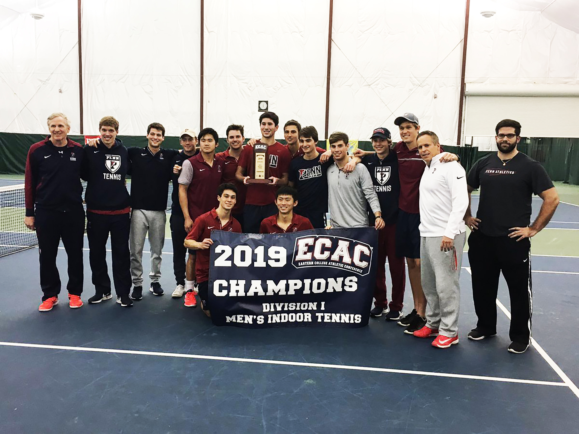Men's tennis players and coaches pose with the 2019 ECAC Championship banner.