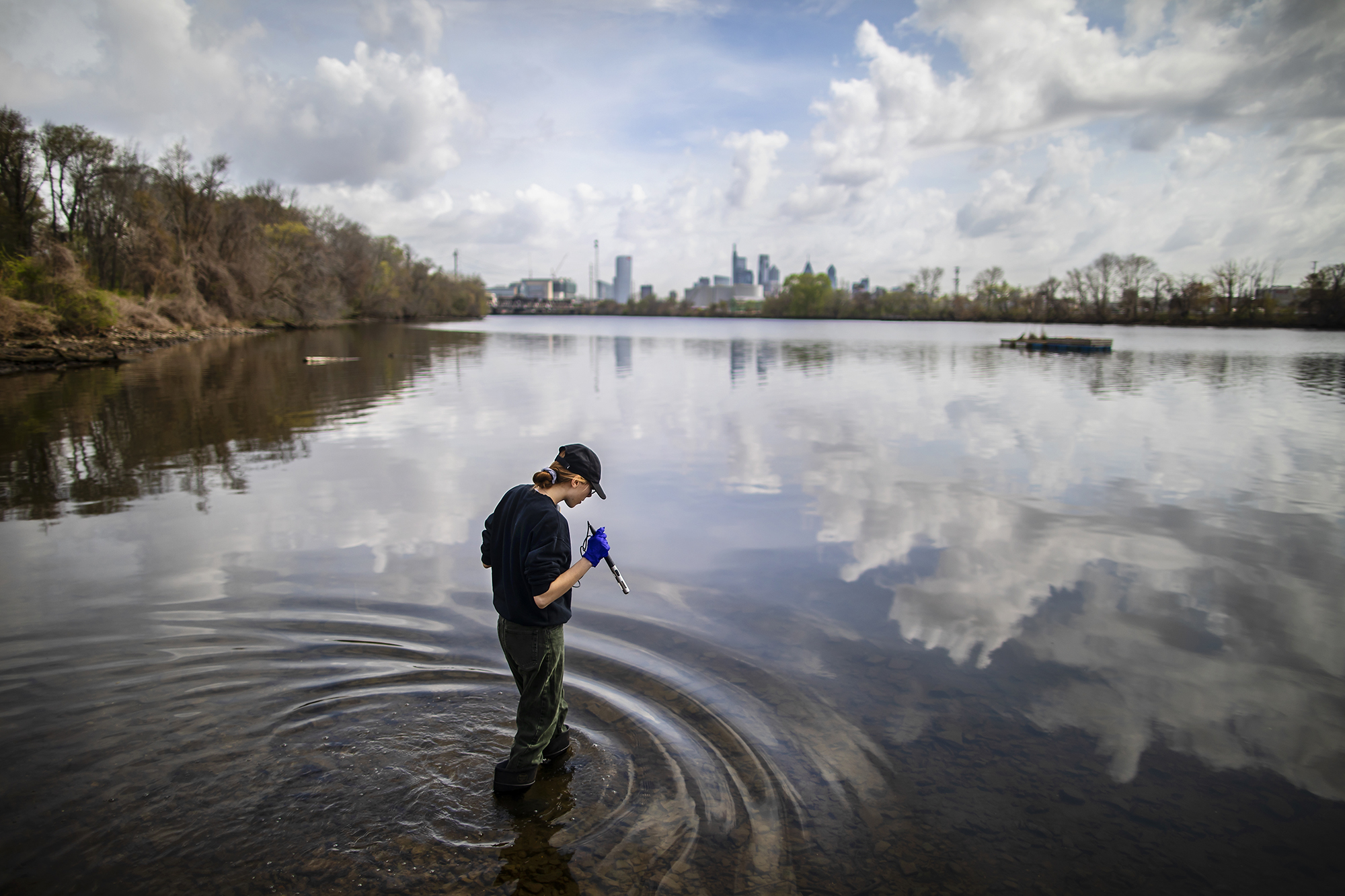 With the Philadelphia skyline in the background, a student in waders and a baseball cap holds a piece of scientific equipment while standing in the Schuylkill River and looking down