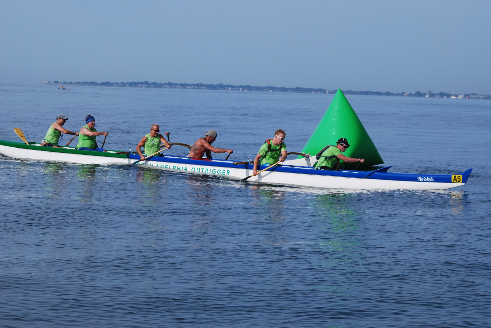 Six people in a canoe labeled "Philadelphia outrigger" turn around a large triangular green buoy