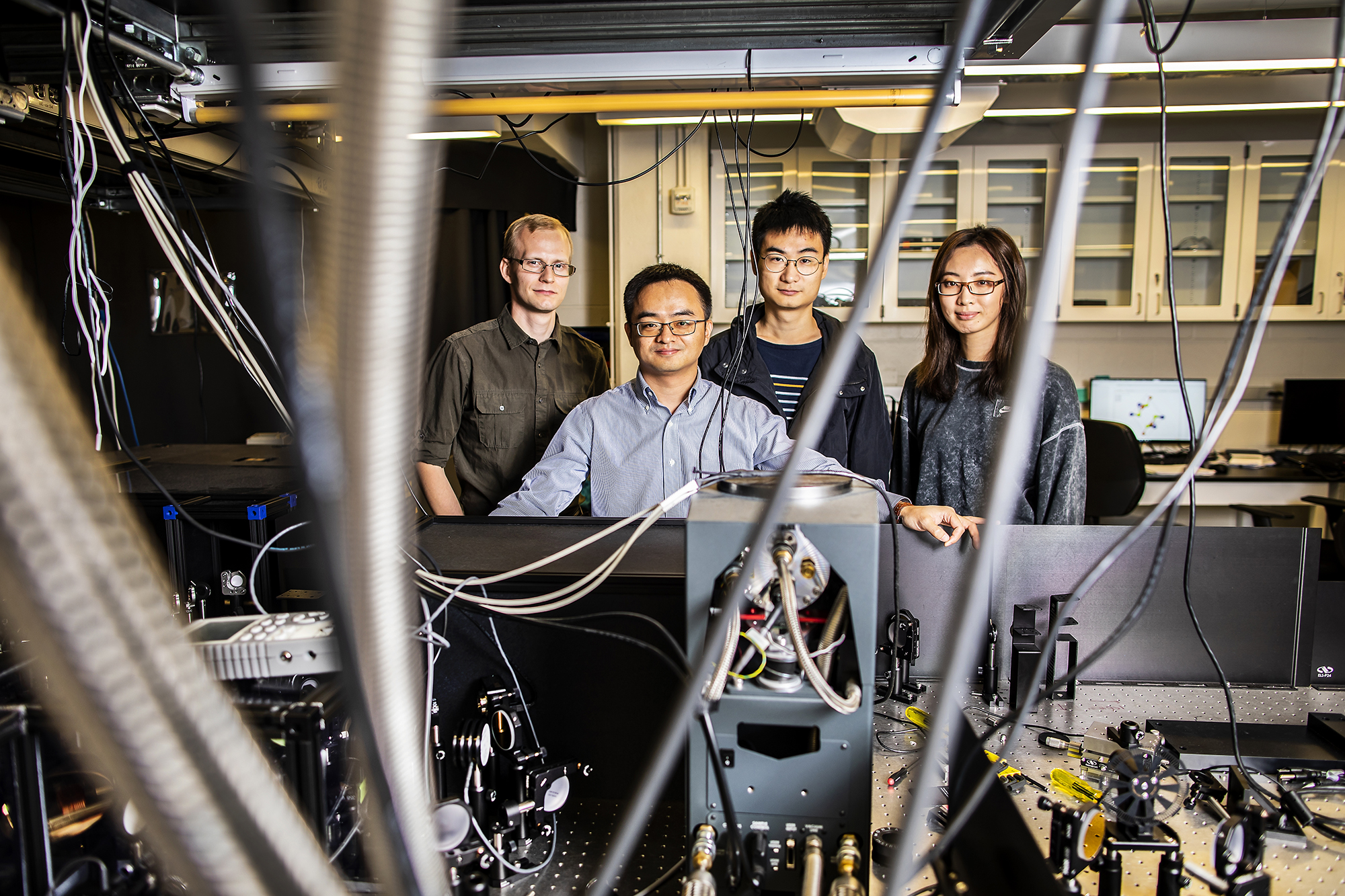 four people standing in front of an optics table behind a tangle of wires in a lab in 