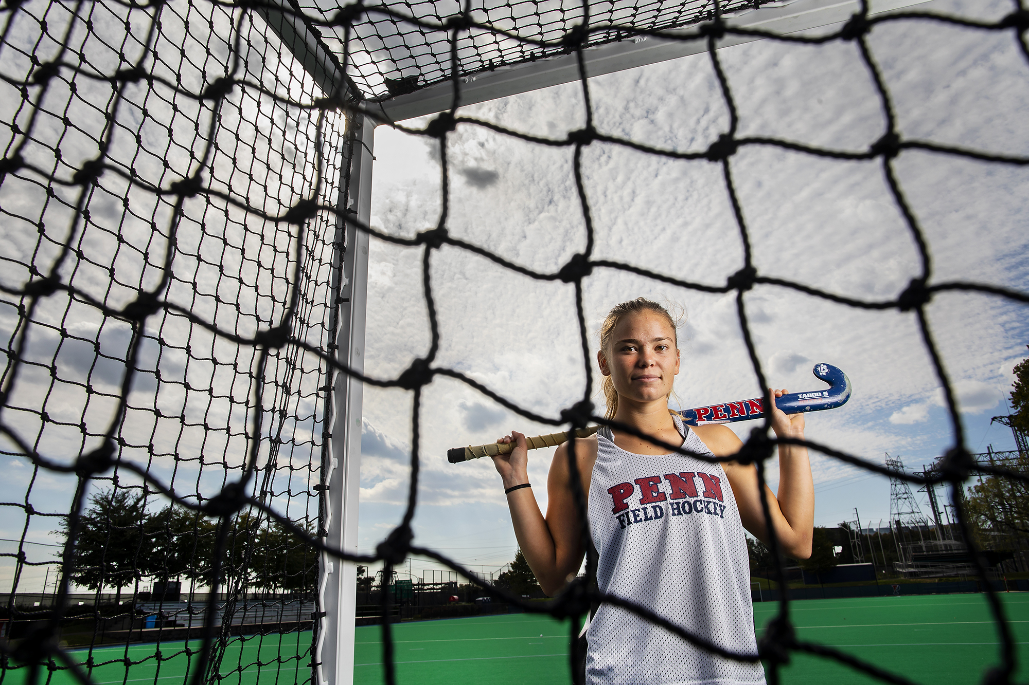 Alexa Schneck of the field hockey team poses on the field hockey field in a net with her stick across her shoulders.