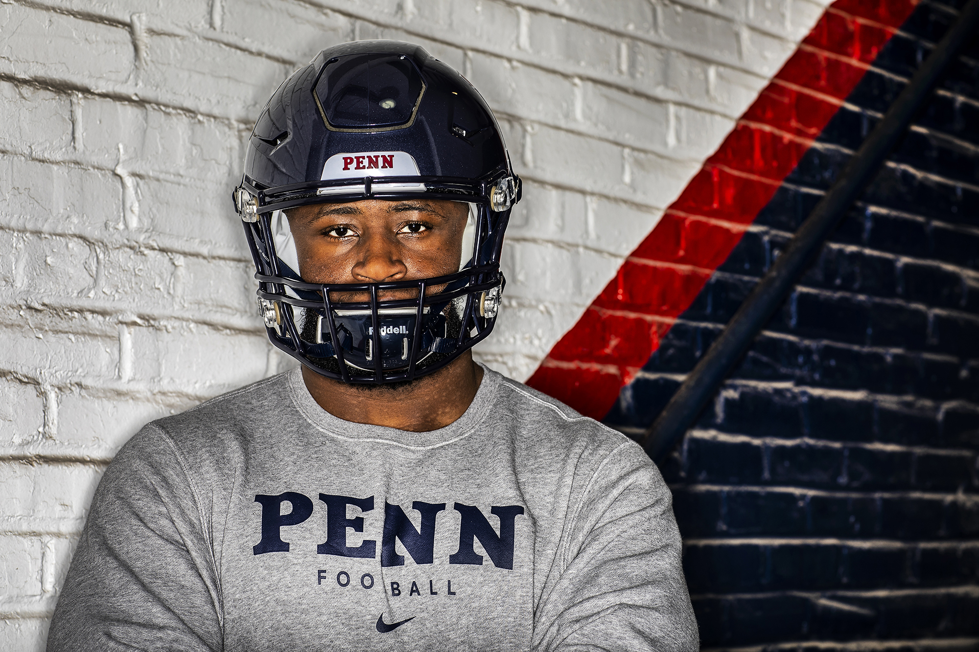 Prince Emili poses wearing his football helmet and a Penn sweatshirt near a red and blue wall at Franklin Field.