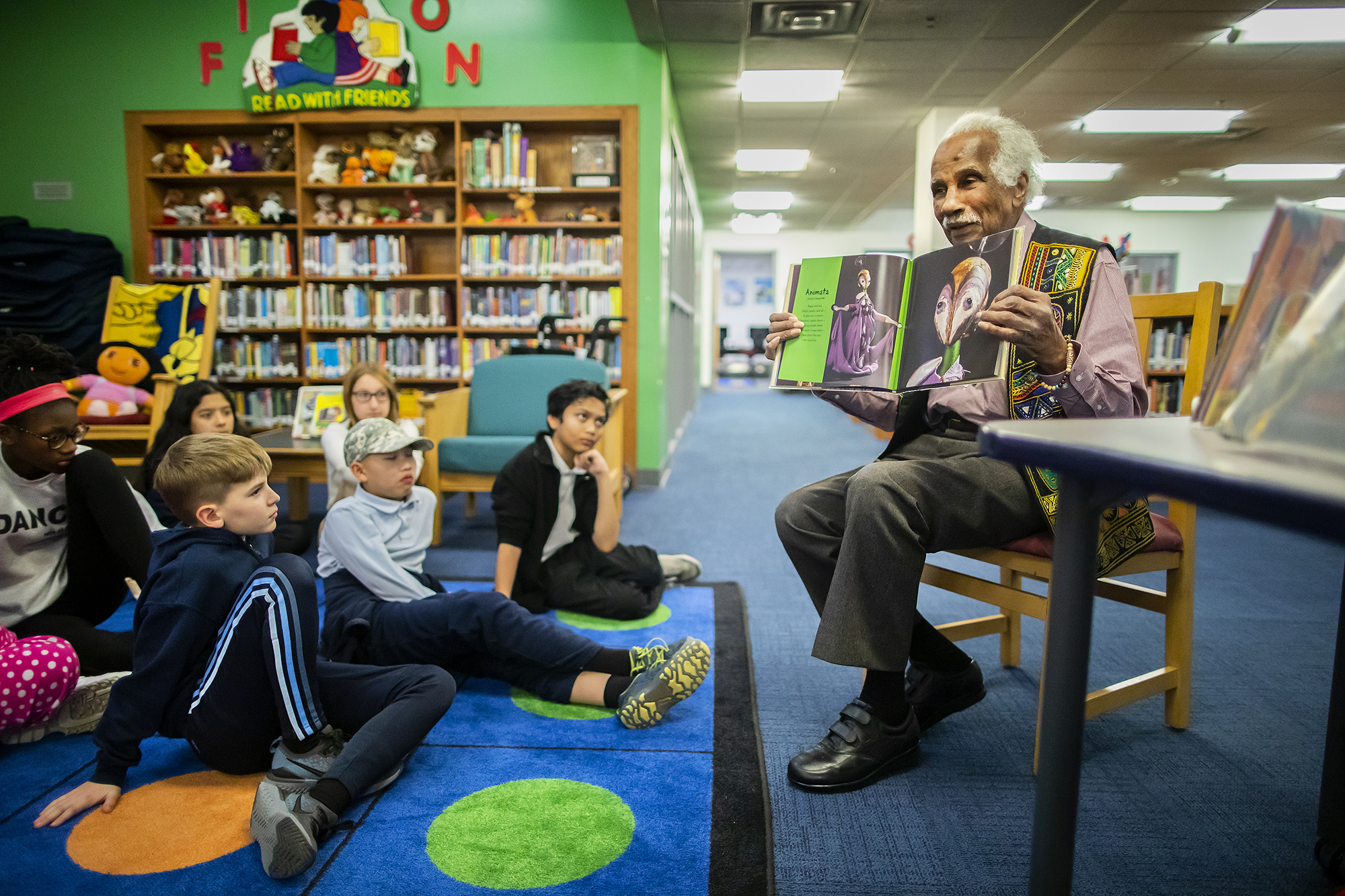 Ashley Bryan holds up one of his illustrated children's books to show a number of elementary students who are seated on the floor in front of him in the Penn Alexander school library.