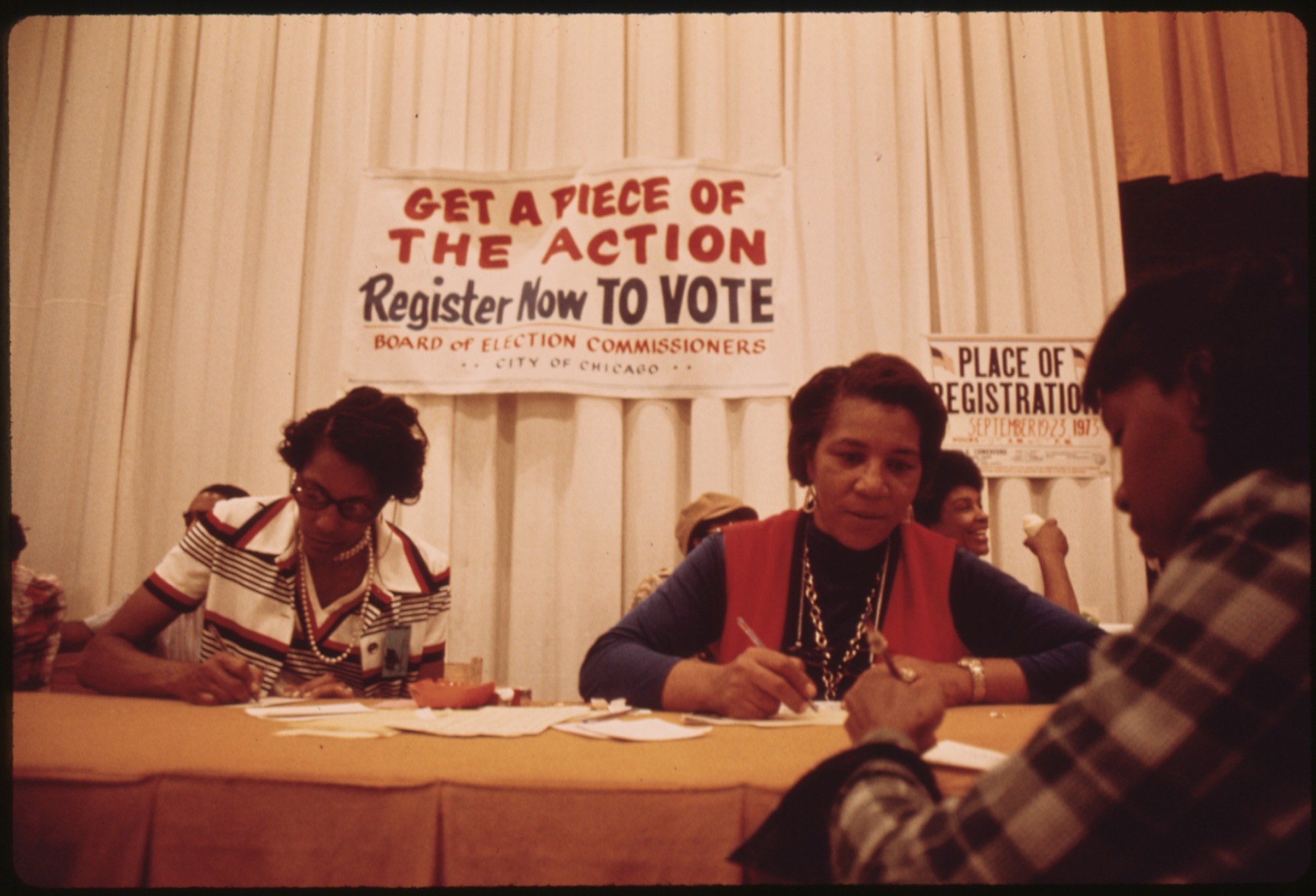 Two people sign papers registering a third person to vote at the Black Expo in 1973, behind the table a sign reads "Get a piece of the action Register now TO VOTE"