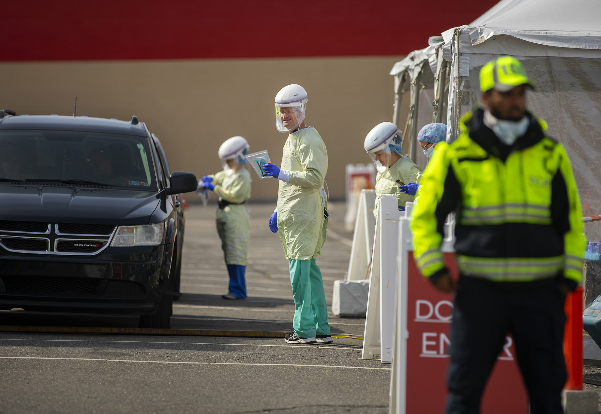 A car in line at drive through testing at the COVID-19 pop up testing site, with four medical personnel outside car in full protective gear and a security guard wearing a medical mask standing at the entrance