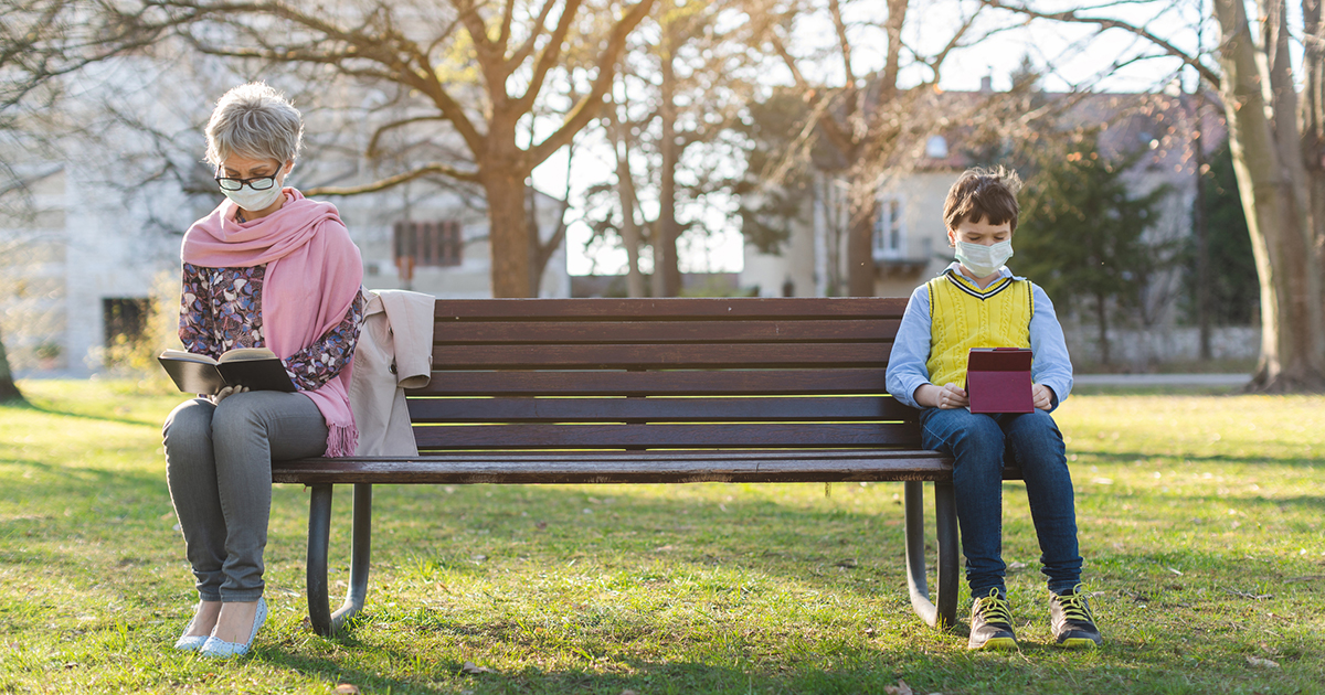A grandparent and a child sit on a park bench in daylight, both wearing masks and sitting six feet apart.