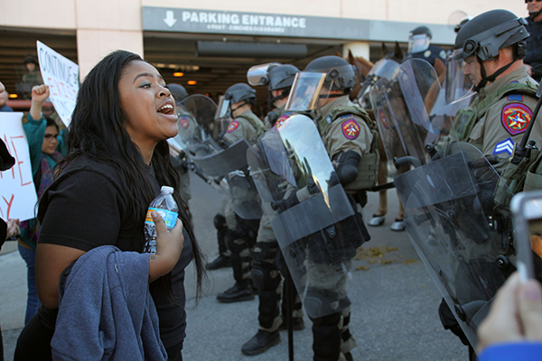 African American person in a crowd at a demonstration confronts a line of state police with riot gear.