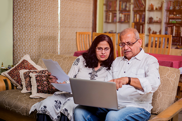 Older couple sitting on a couch looking at a laptop, one partner holds papers in their hand.