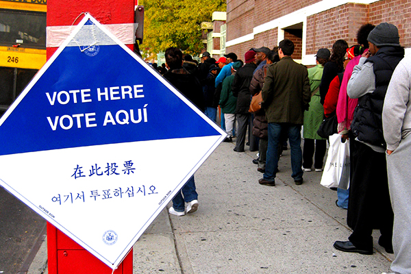 line of people at a polling place with a sign out front reading VOTE HERE/VOTE AQUI and also in Mandarin and Cantonese.