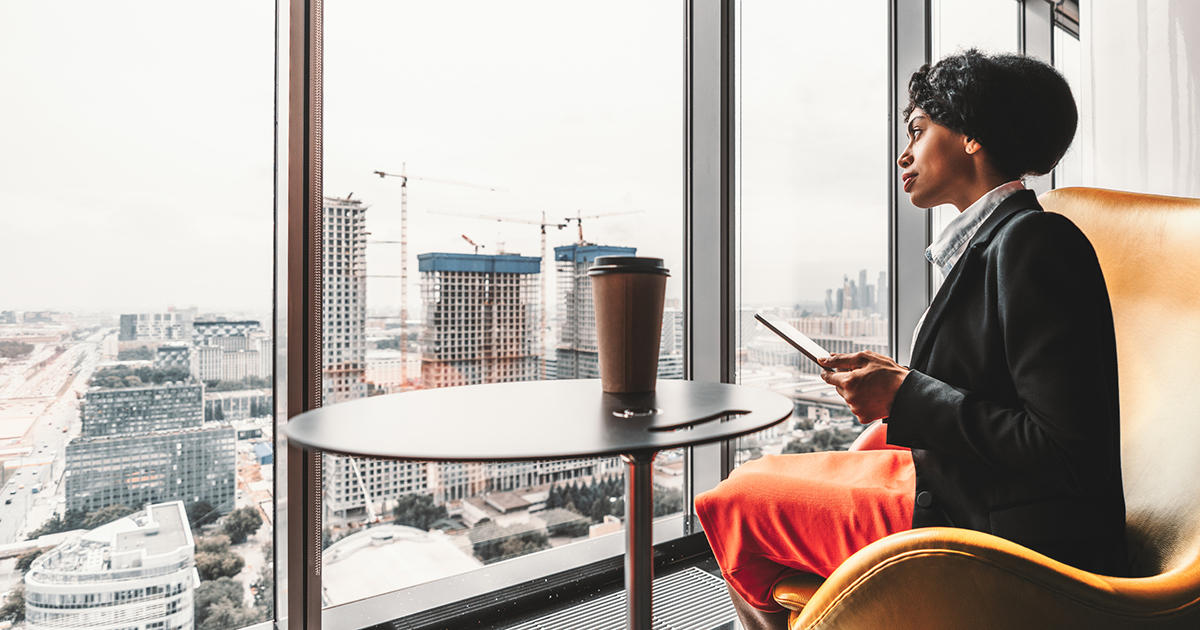 Professional African American person using a digital tablet looking out the windows on the top floor of a city office skyscraper.