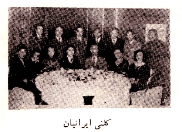 A group of 14 people line up behind a round dinner table at a restaurant to pose for photo