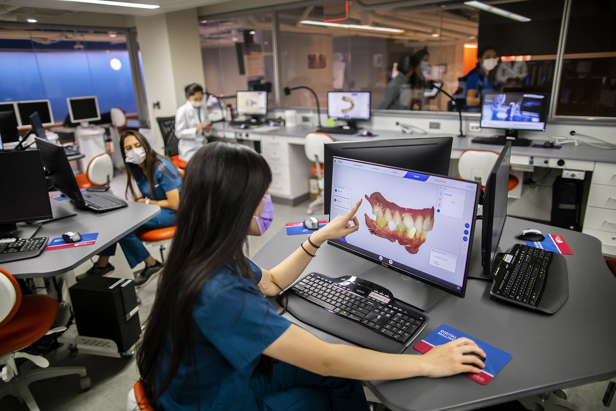 A person in a dental computer lab sits at a computer and points to a virtual model of a mouth and teeth