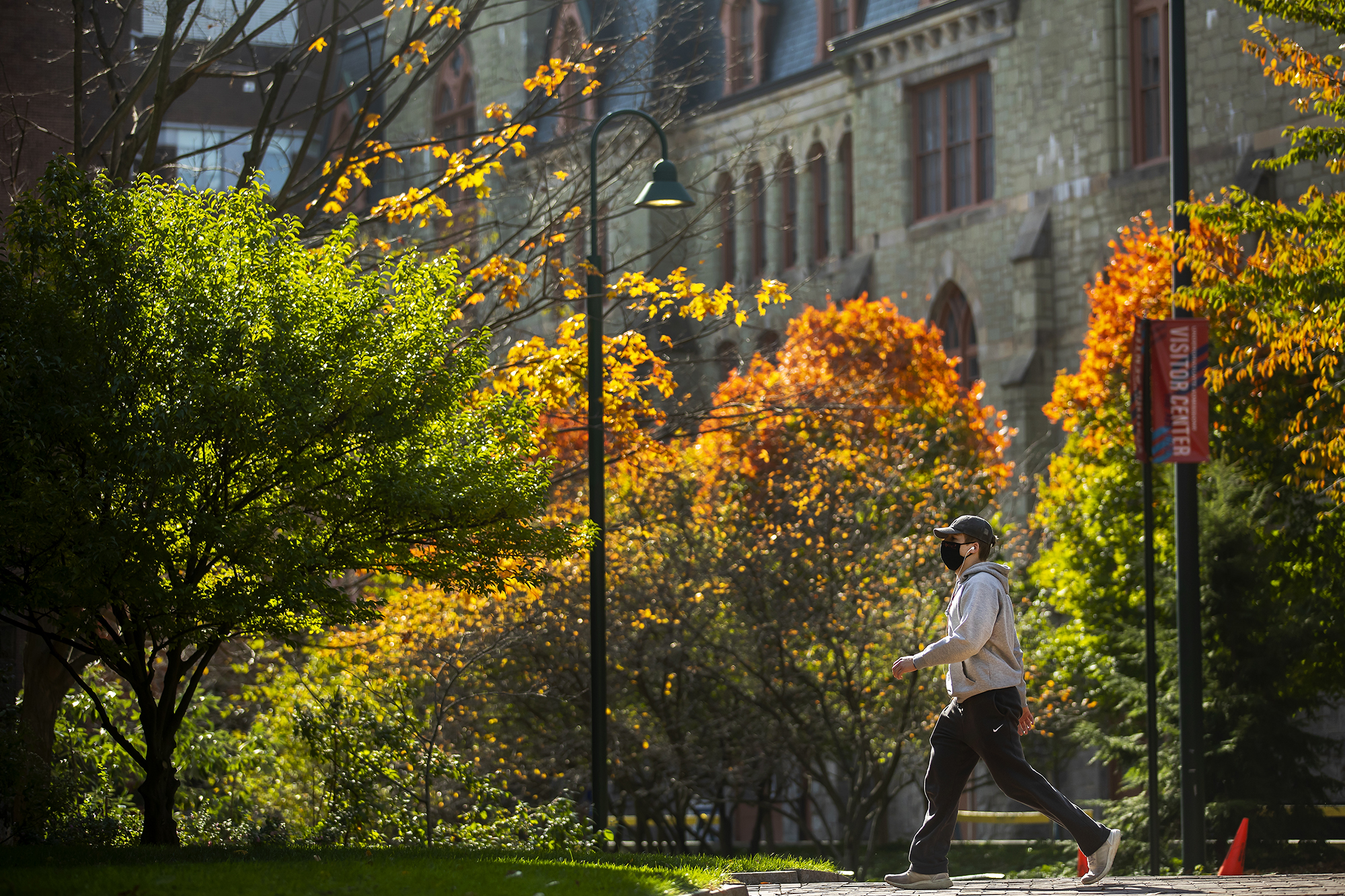 Person wearing a face mask walking on Locust Walk with College Hall in the background surrounded by colorful fall foliage.