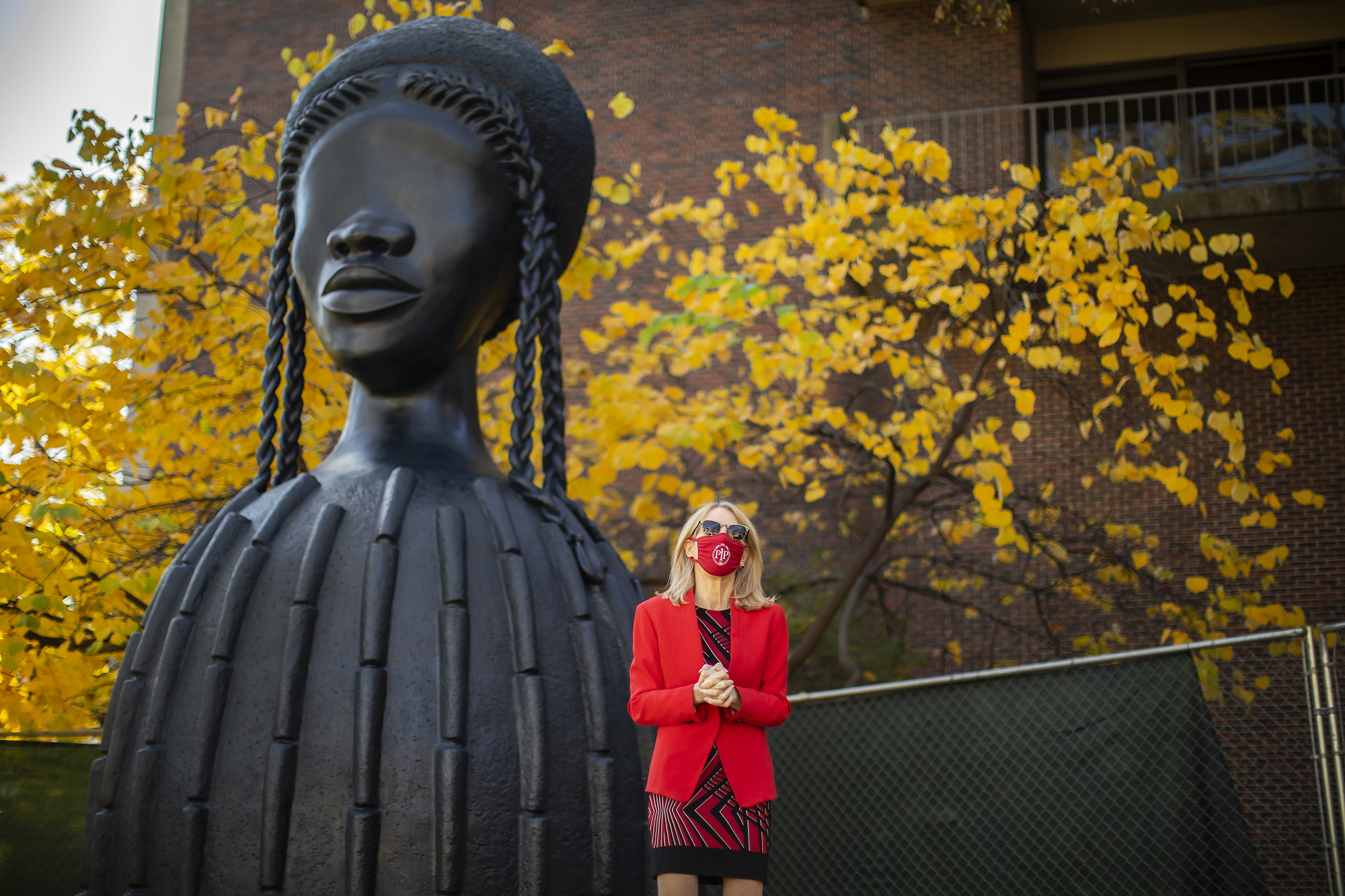 Amy Gutmann wearing a mask and sunglasses stands in front of the bronze sculpture “Brick House”