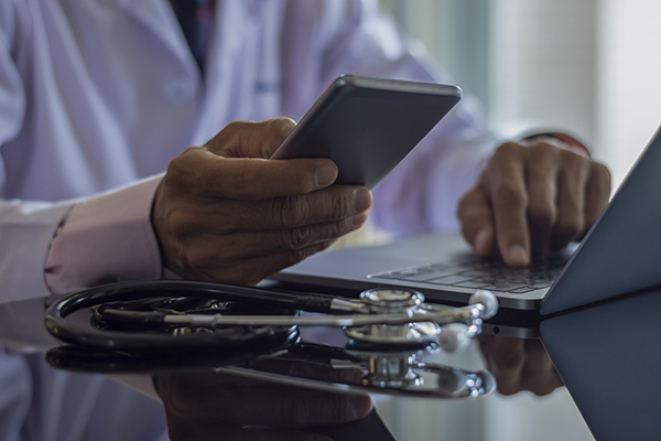 Closeup of doctor’s hands holding a smartphone over an open laptop beside a stethoscope.