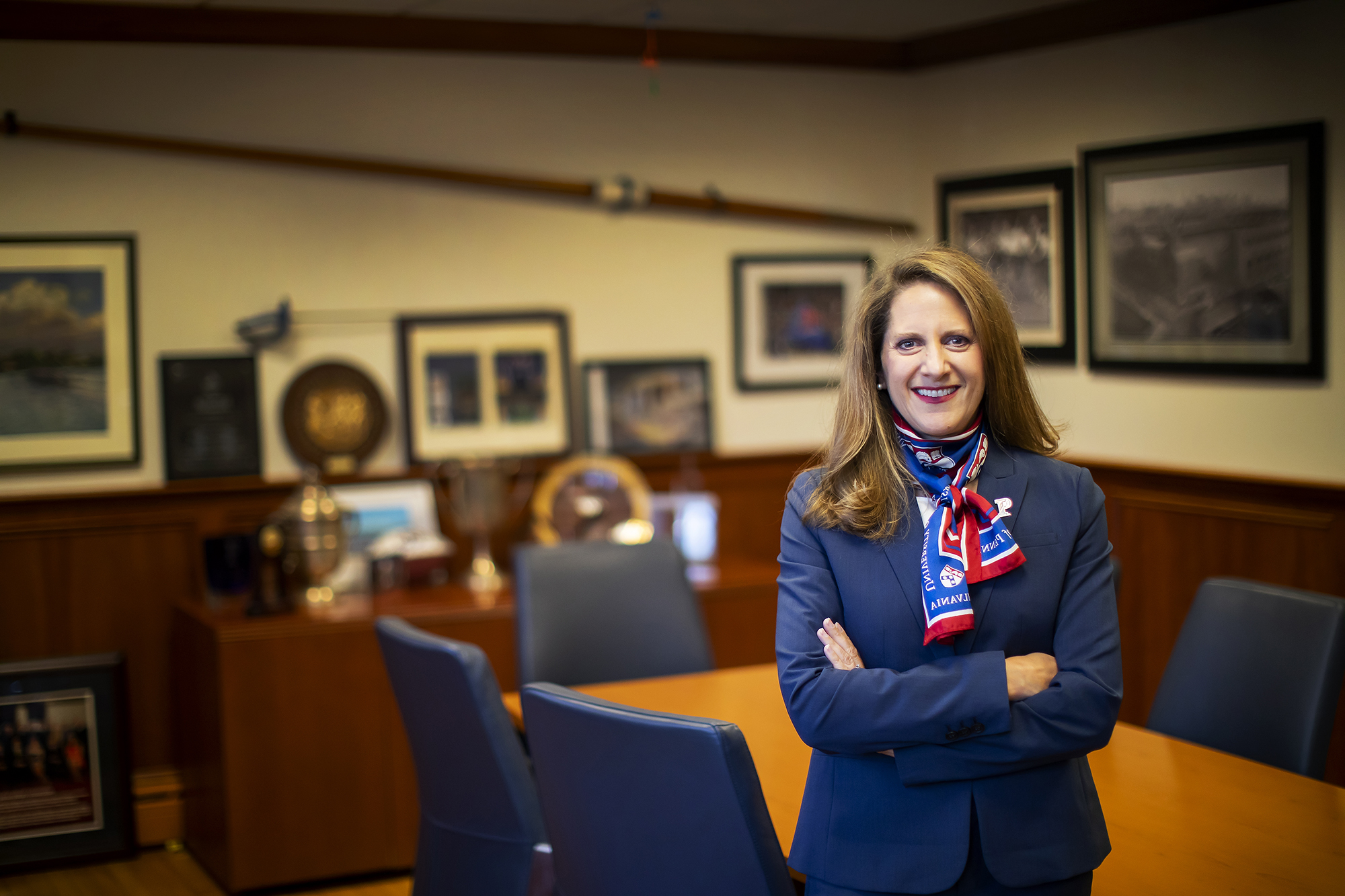 AD M. Grace Calhoun stands with her arms folder while wearing a blue suit and Penn scarf in her office.