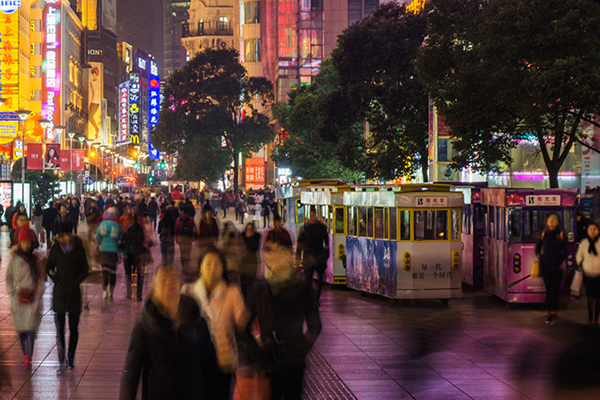 Busy sidewalk in urban China at night with crowds walking past commercial businesses.