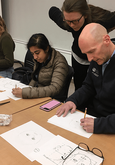 Two students drawing medical imagery in a PSOM class on medical drawing, with an instructor standing over them, observing.