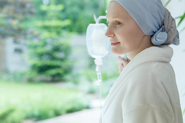 Cancer patient with IV drip and scarf in their hair looks out the window.