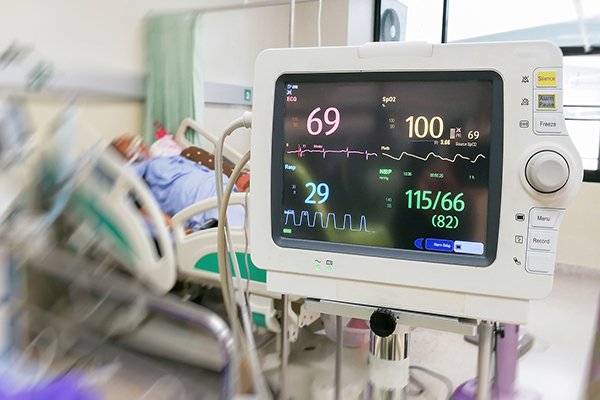 Heart monitor in a hospital room, a patient in a hospital bed behind it.