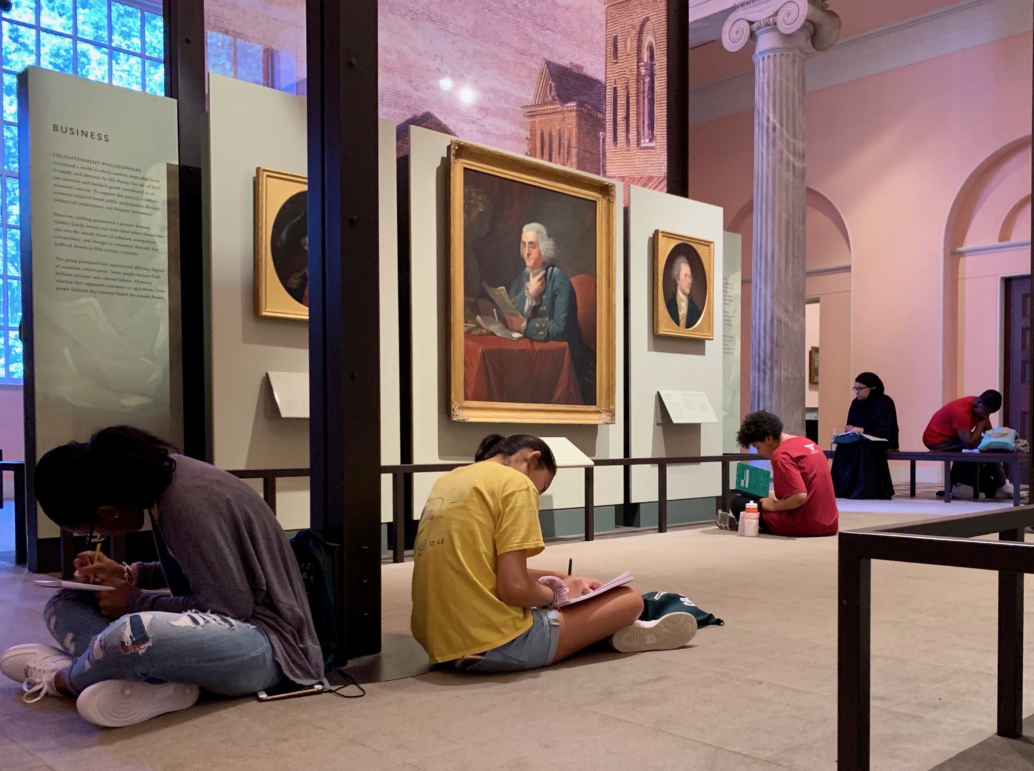 Students seated on ground write in notebooks in a museum