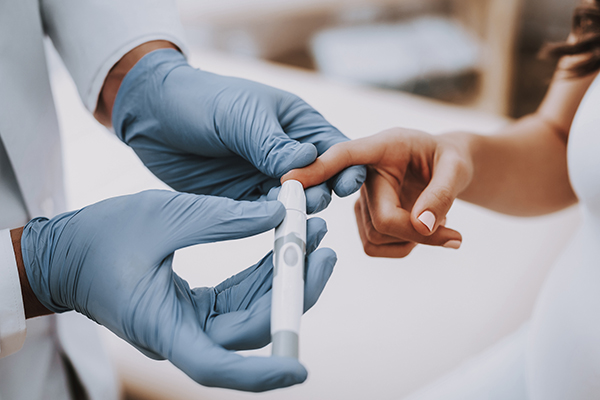 Gloved hand administering a blood sugar finger test to a hand of an African American person.