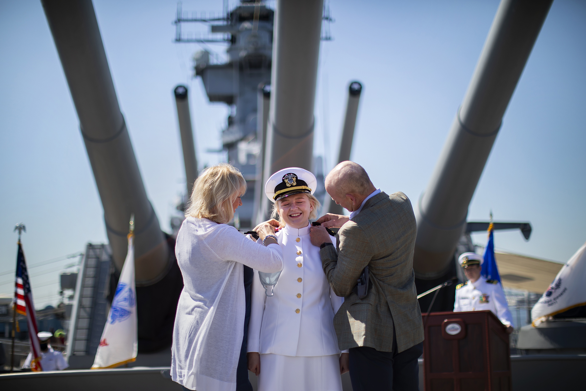 Person in uniform smiles while parents put medals on their uniform aboard the USS New Jersey.