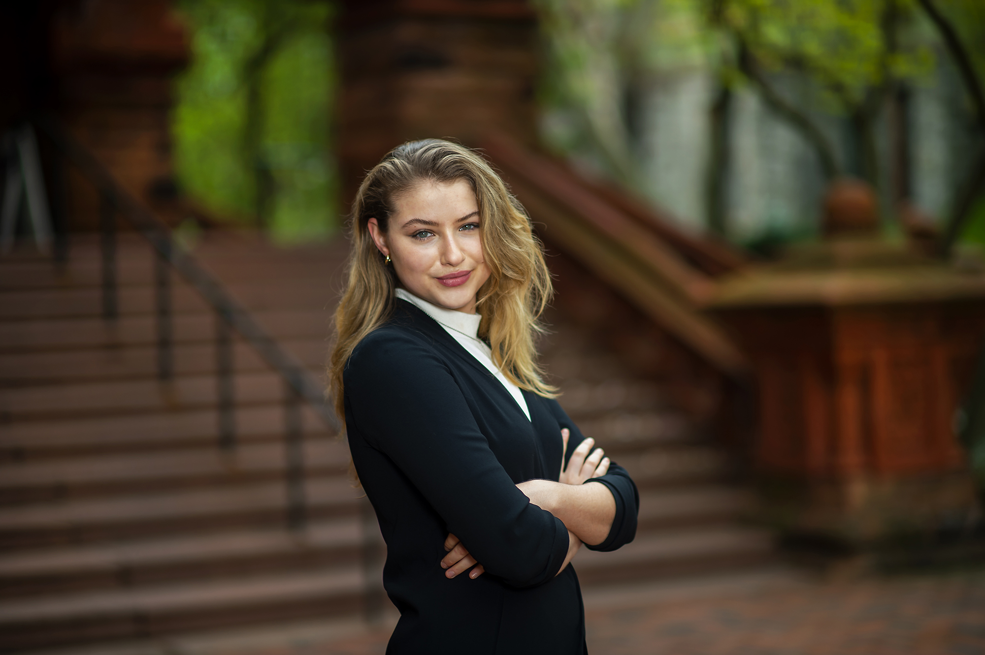 Woman with blond hair in a black suit with white mock turtleneck stands with arms crossed in front of a brown staircase and looks into the camera