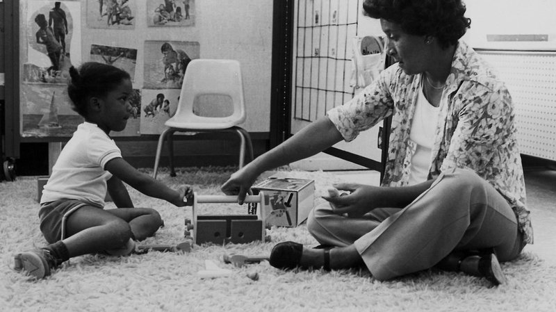 A black-and-white image of an adult sitting on a carpeted floor with a young child. Between them are two toys. Behind them is a plastic chair and some images on the wall.