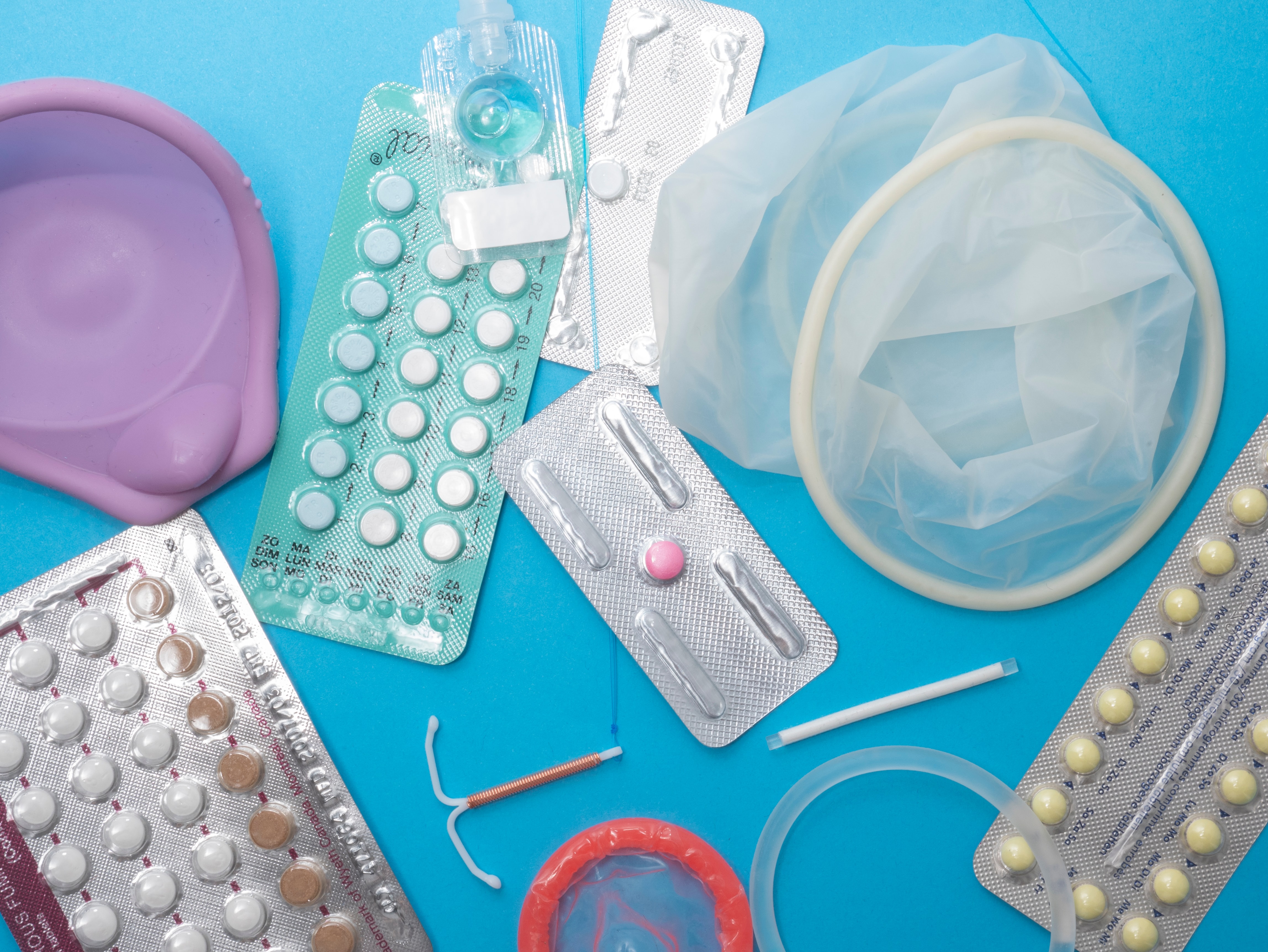 A flat lay of bIrth control pills, IUDs, condoms, and other contraceptives against a blue background