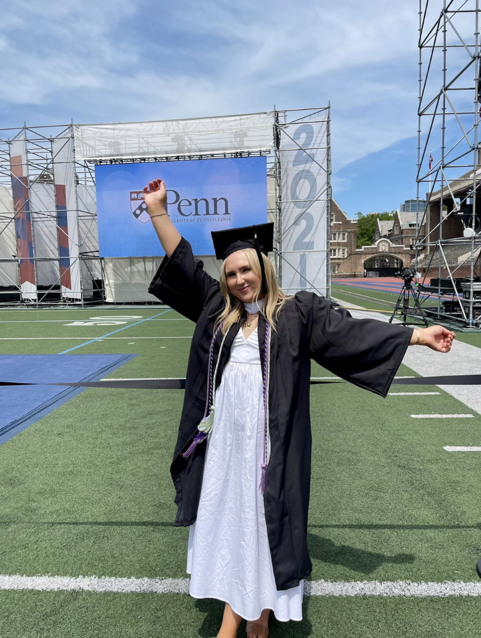 student in graduation gown standing on Franklin Field with Penn 2021 sign