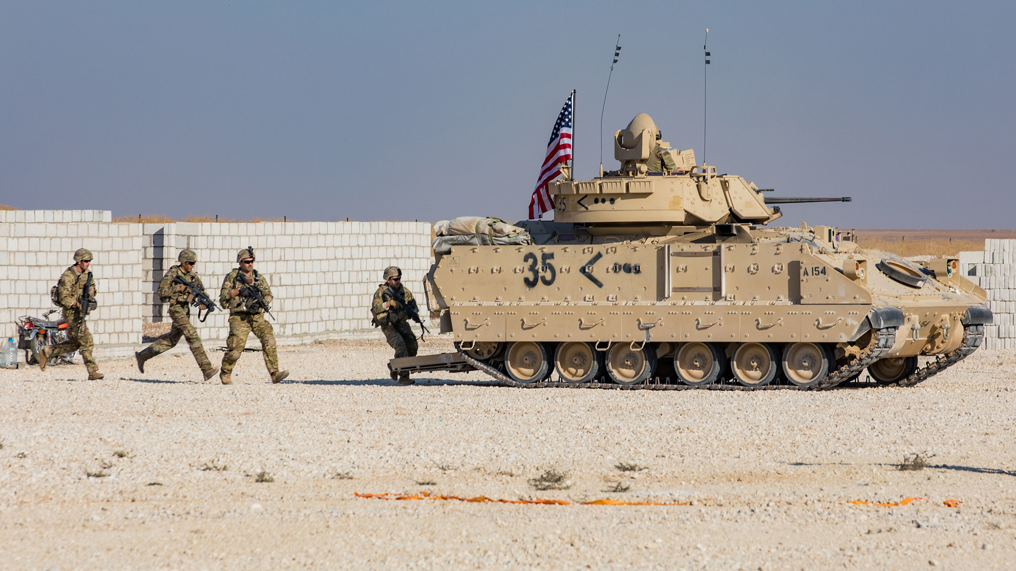 US soldiers holding guns are running behind a tank with an American flag on top in sand in Syria 