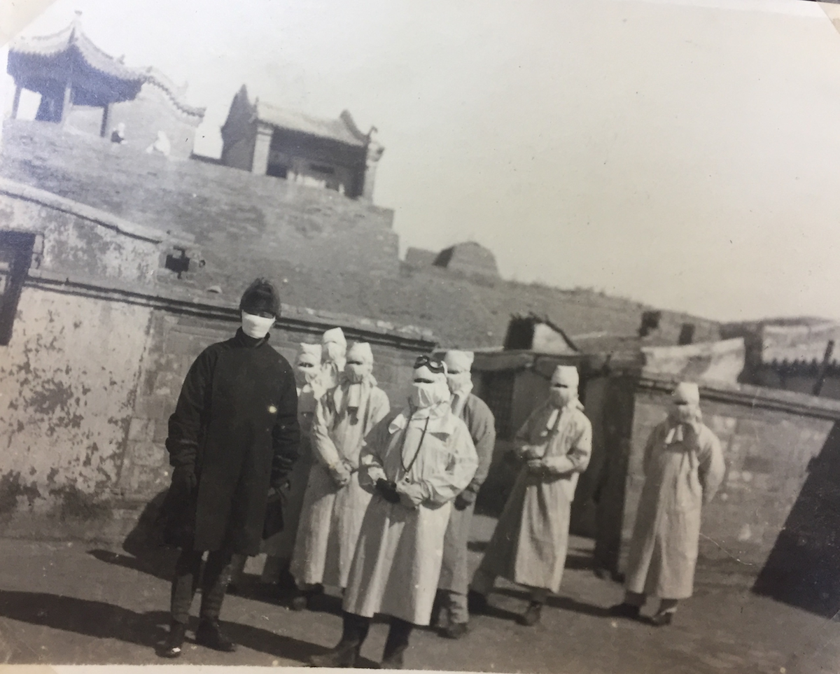 People wearing medical gear and medical face masks stand in front of a Chinese temple in 1918