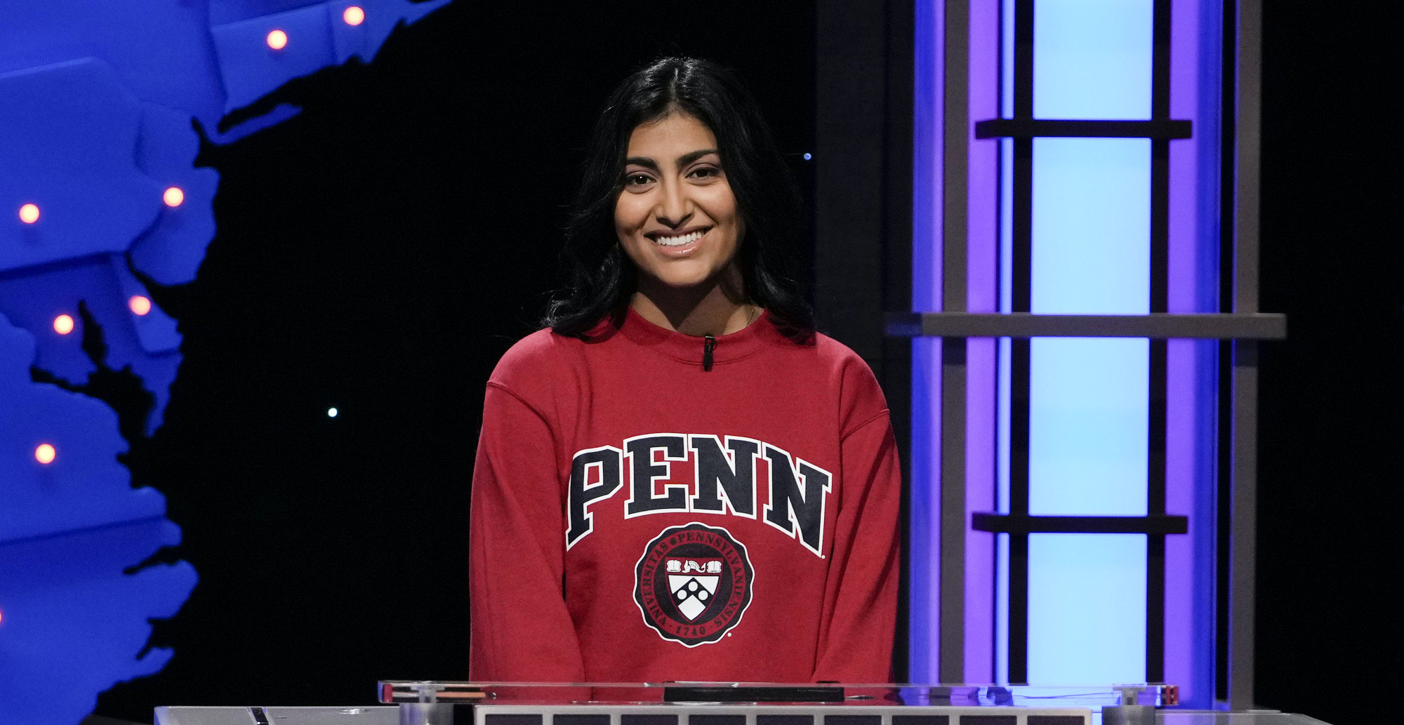 a smiling student wearing a Penn sweatshirt stands in front of a game show platform