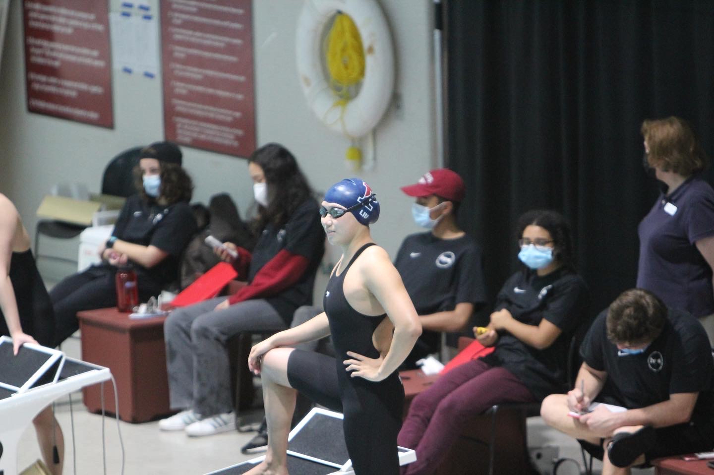A woman stands with one foot on the starting block before a swim meet. Onlookers are masked behind her.