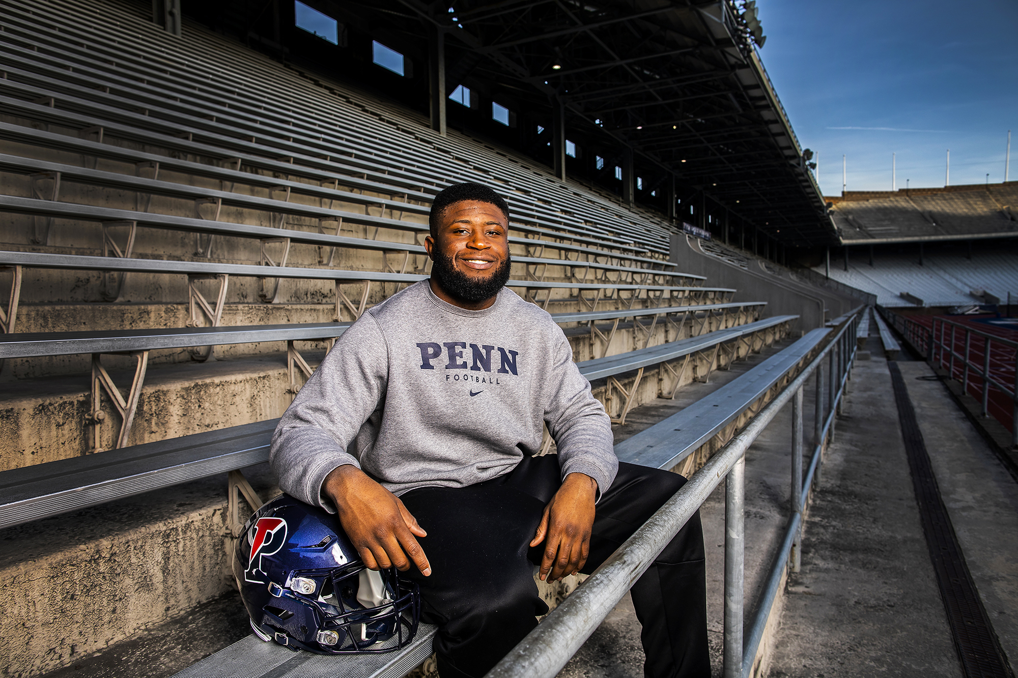 Emili sits in the bleachers at Franklin Field with his arm on a Penn helmet.