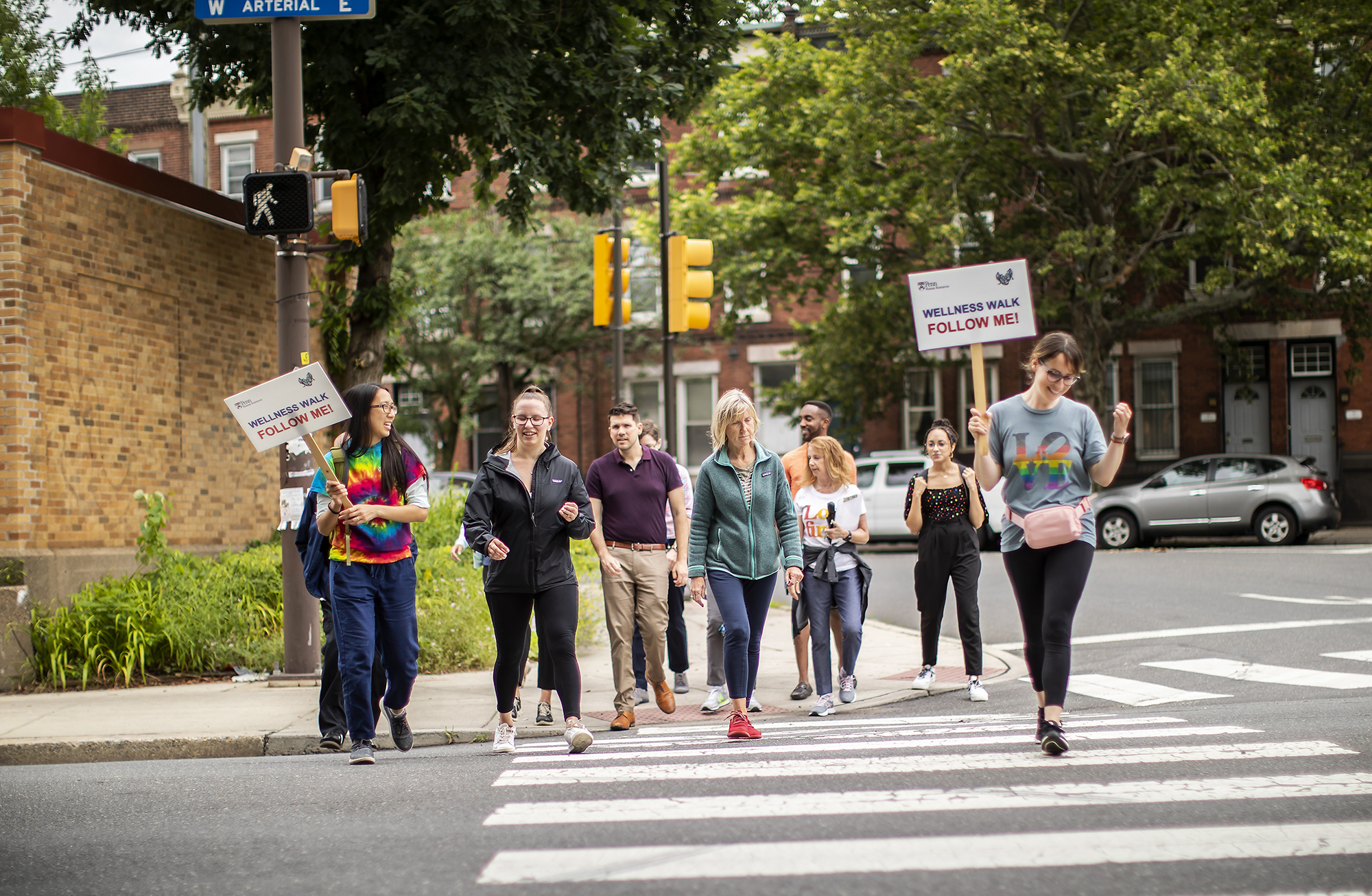 A group of people crossing the street. Signs read: Wellness Walk, follow me!