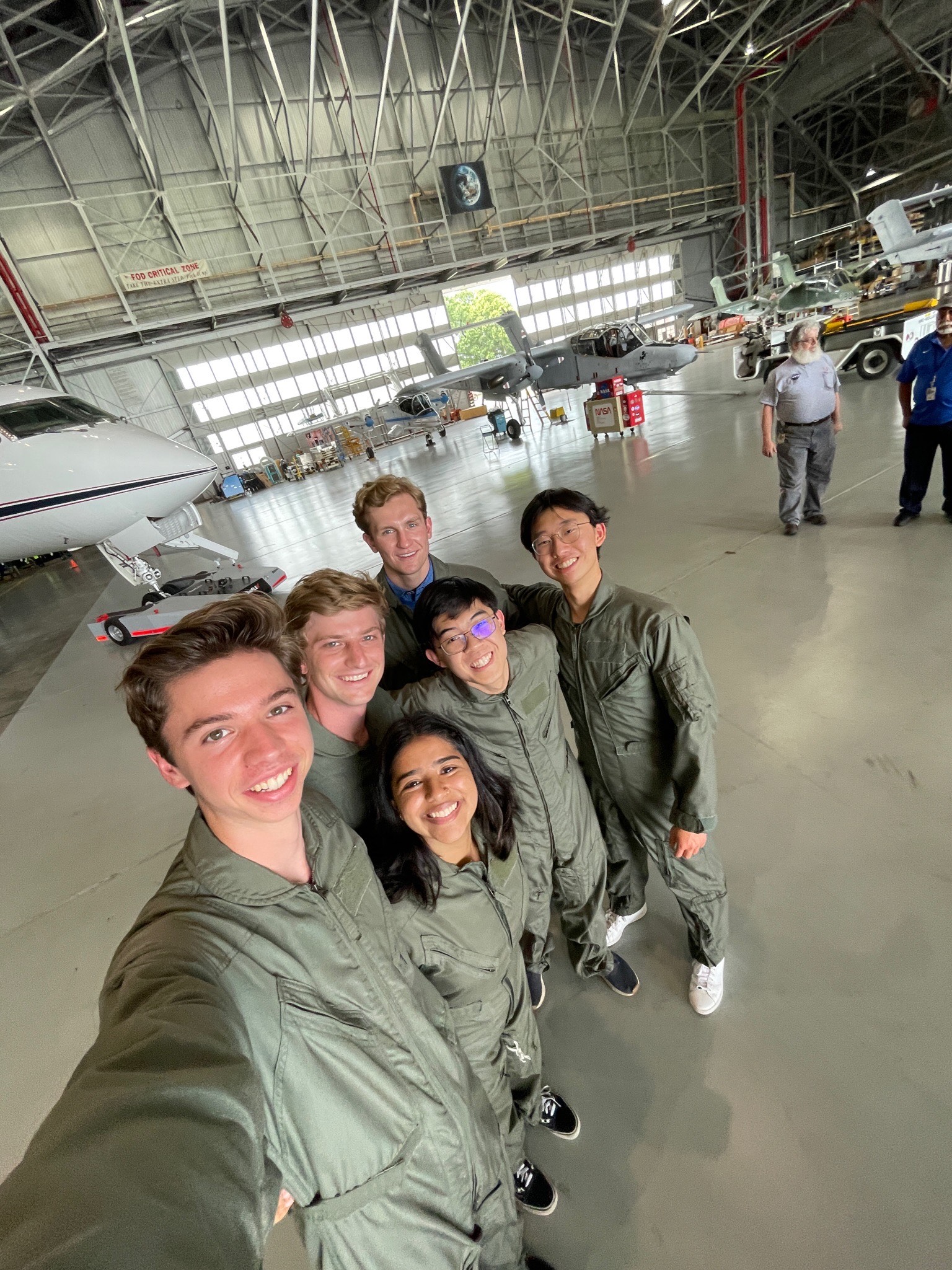 group of six students pose wearing flight suits in an airplane hangar with planes in background