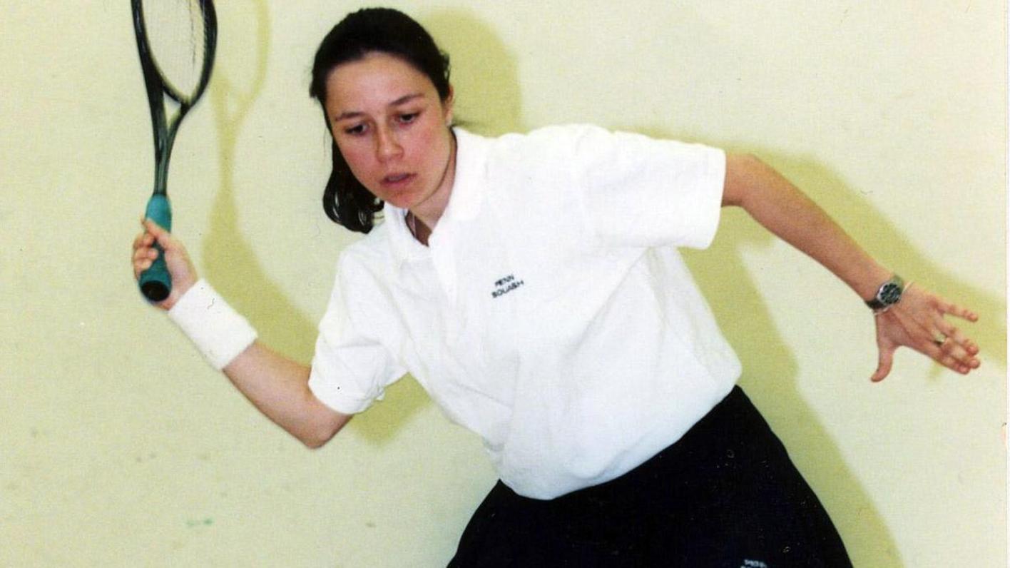 Jessica DiMauro demonstrates her swing in front of a wall in the squash court, bending right at the waist, holding a racquet up with the right hand and reaching out to the side with the left hand. 
