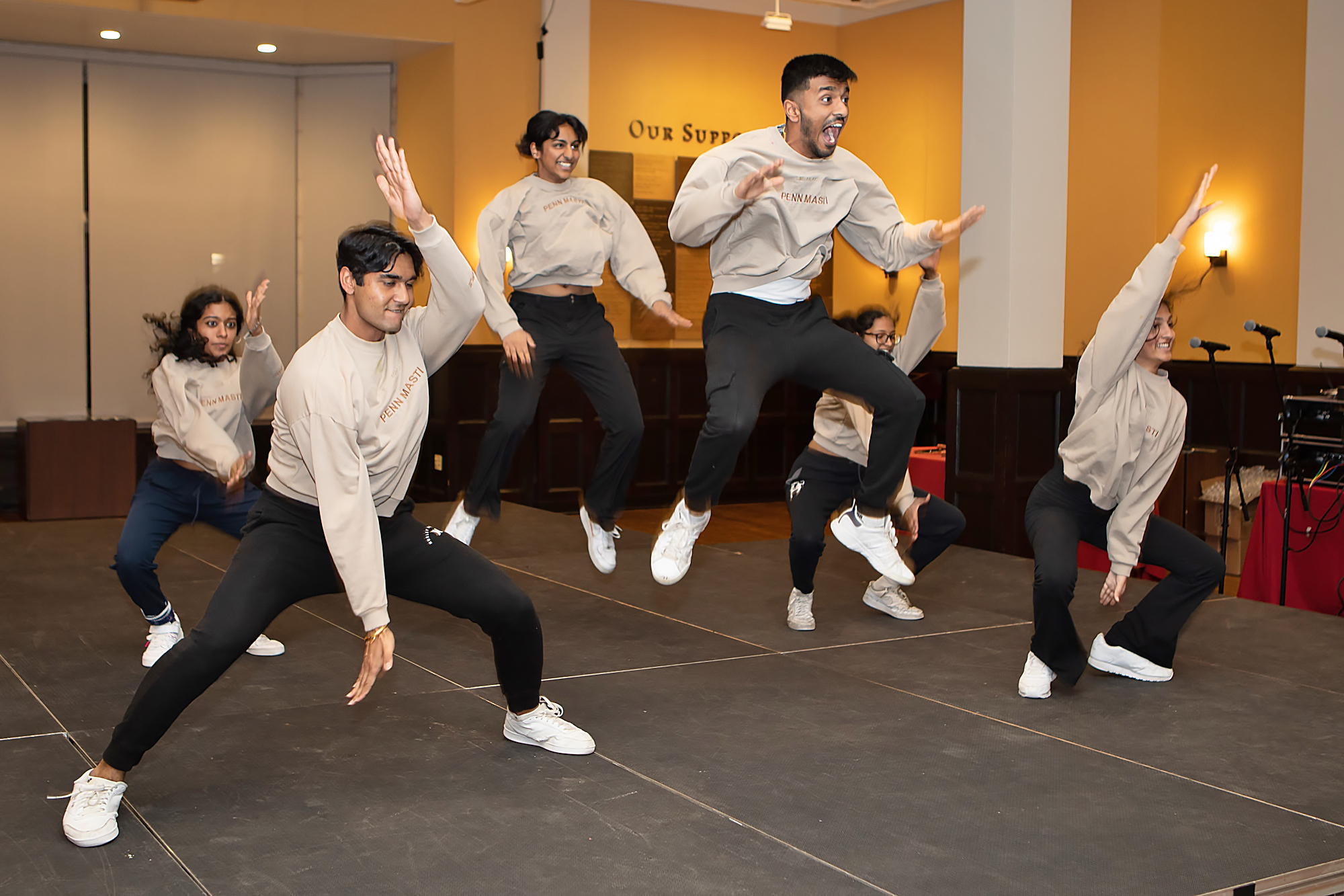 Six dancers in uniform beige and black sweatsuits squat and jump on stage