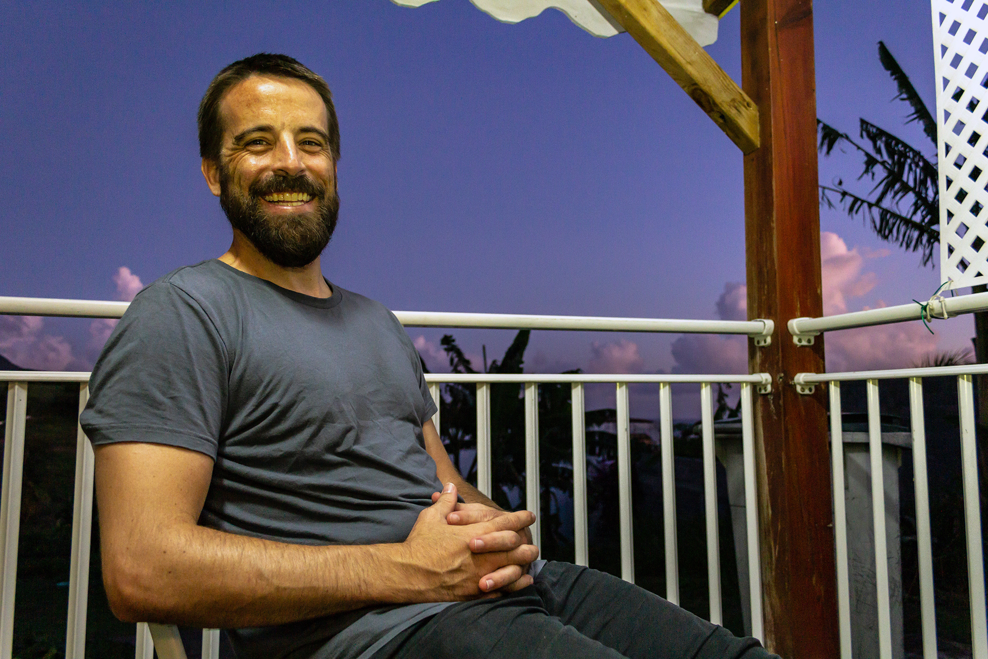 Man with beard smiles at camera sitting on a balcony with a sunset and palm trees behind him, hands folded on his lap.