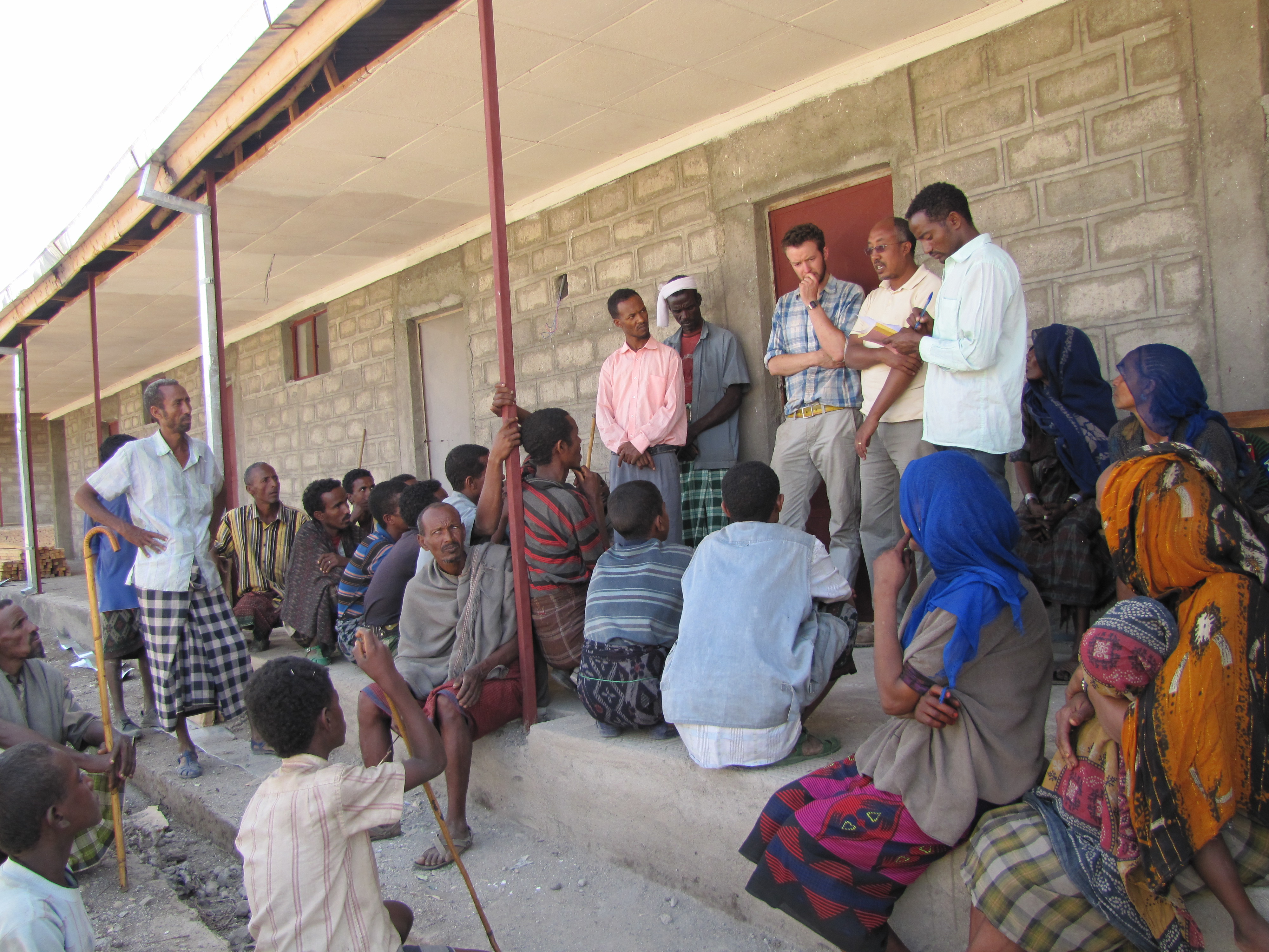 A few people stand and speak to a group of people seated on and around the porch of a building
