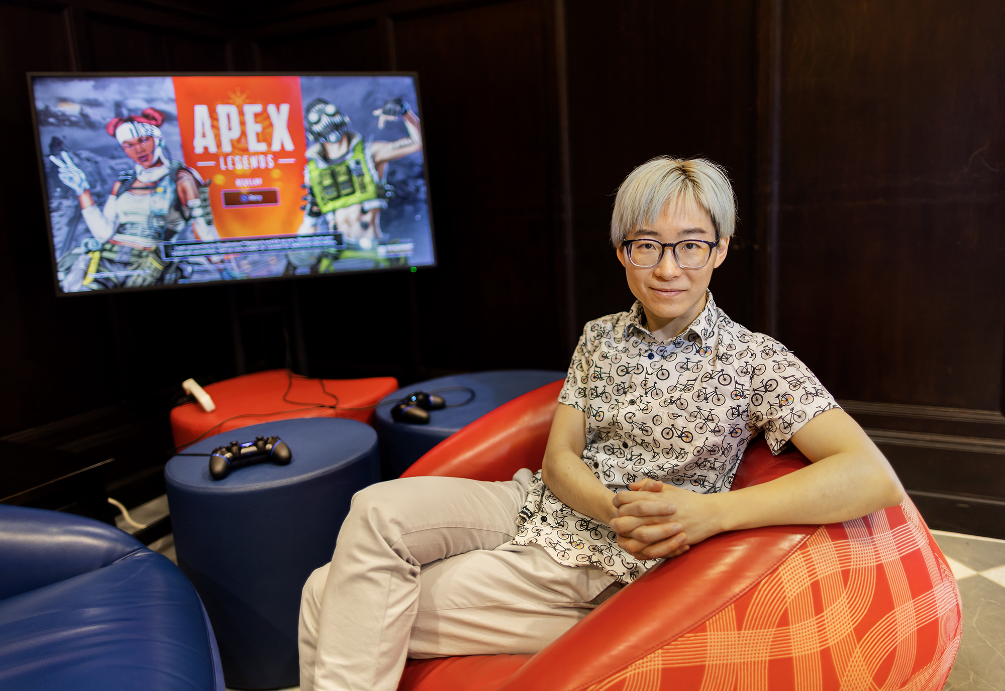 Mengyang Zhao rests on a bean bag chair with a video game that reads 'Apex Legends' in the background