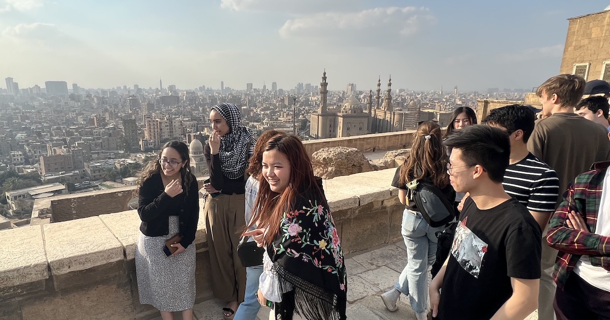 A group of students take pictures on a rooftop deck. The city of Cairo, with its skyscrapers and minarets, are visible in the background