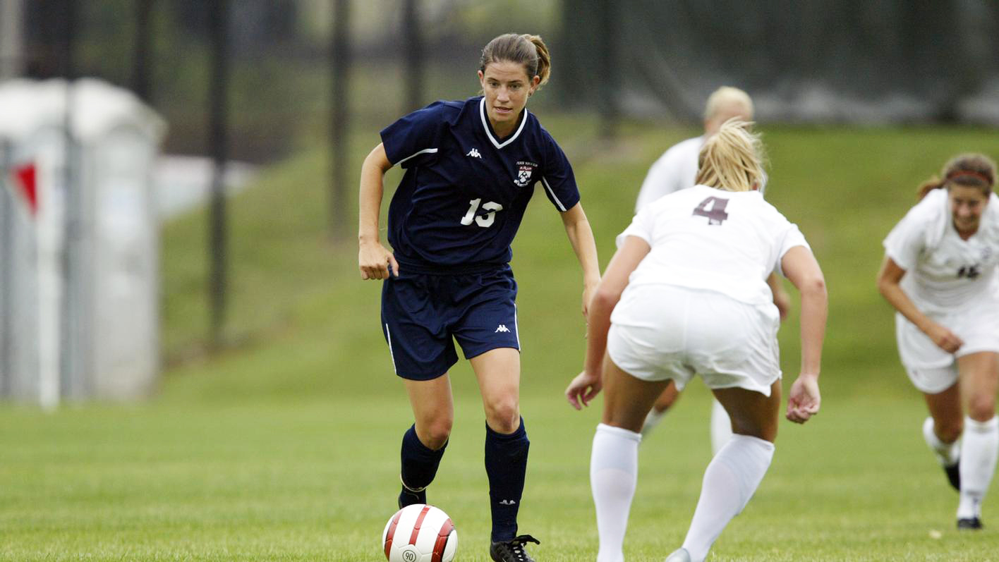 Katie Cross, wearing a blue Penn jersey and shorts, dribbles the ball past a defender.