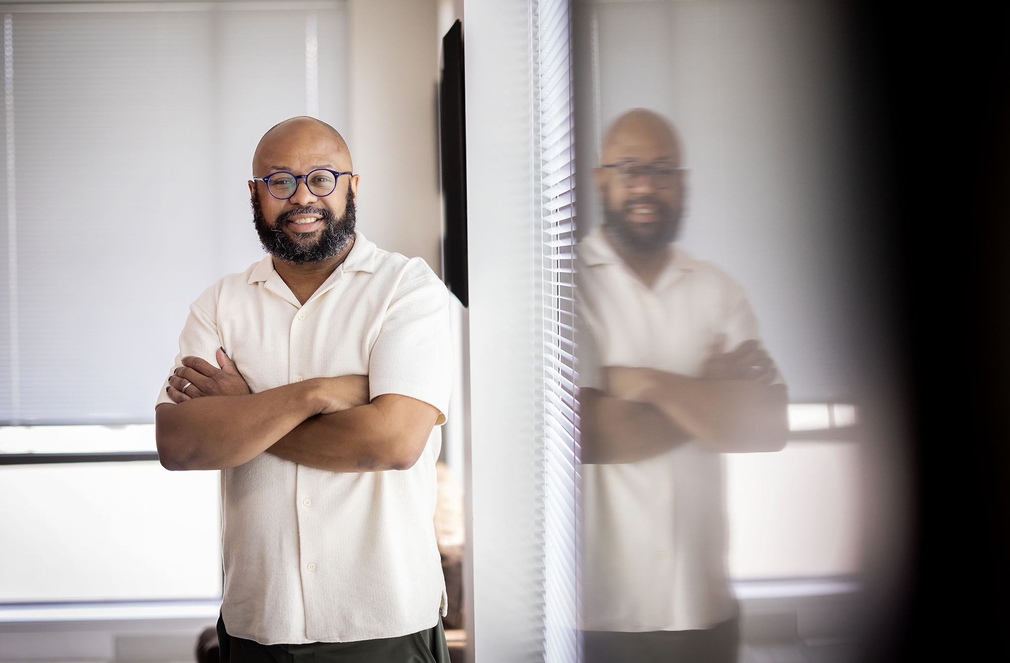Desmond Patton stands with arms crossed in front of a window, which mirrors his image